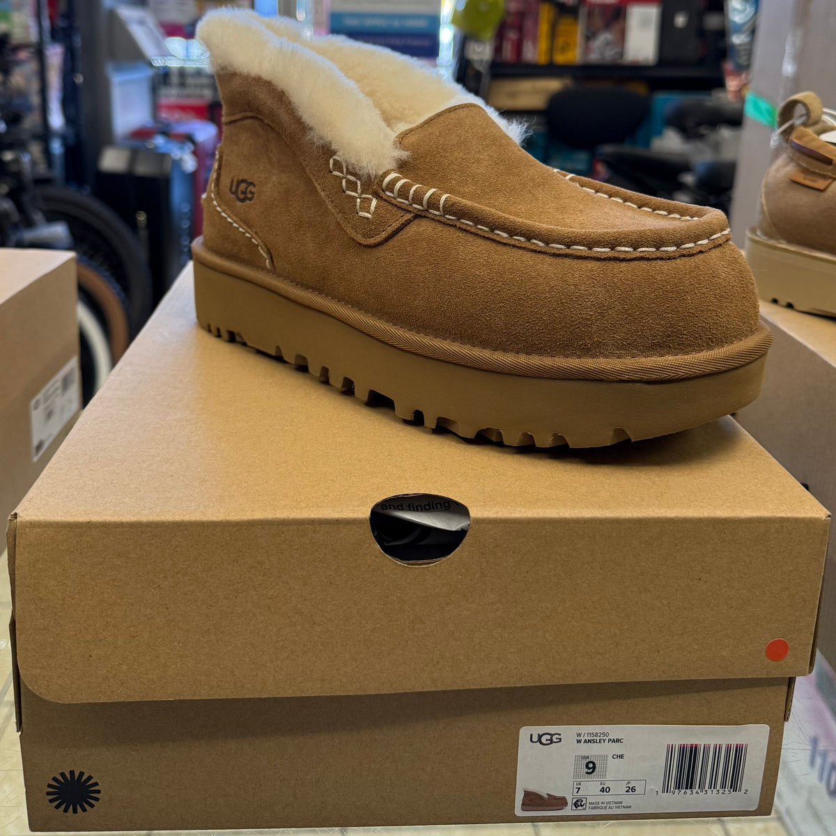 Tan suede UGG W Ansley Parc slipper with white fleece lining, decorative stitching, and thick rubber sole, displayed atop its labeled cardboard box showing size 9 US, made in Vietnam.